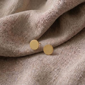 Puede incluir: Un par de pendientes de bot&oacute;n circulares de color dorado. Los pendientes se muestran sobre una tela beige texturizada. El dise&ntilde;o simple y minimalista de los pendientes contrasta con la tela suave y drapeada.