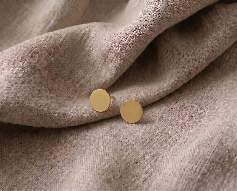 Puede incluir: Un par de pendientes de bot&oacute;n circulares de color dorado. Los pendientes se muestran sobre una tela beige texturizada. El dise&ntilde;o simple y minimalista de los pendientes contrasta con la tela suave y drapeada.