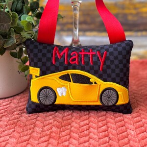 May include: A black and gray checkered tooth fairy pillow with a yellow race car design and the name "Matty" embroidered in red. The car has a tooth graphic on the side. A red ribbon is attached for hanging.
