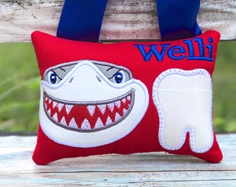 Könnte beinhalten: Ein rotes Kissen mit einem weißen Hai-Gesicht und einem weißen Zahn, die darauf gestickt sind. Das Kissen hat ein blaues Band daran befestigt und der Text "Welli" ist in blauer Farbe auf das Kissen gestickt.