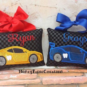 May include: Two tooth fairy pillows with car designs. One is yellow with the name "Ryan" in red script and a red bow. The other is blue with the name "Grayson" in blue script and a blue bow. Both have a black and gray checkered pattern.