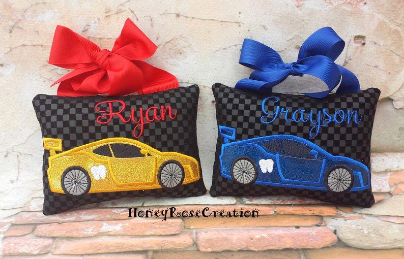 May include: Two tooth fairy pillows with car designs. One is yellow with the name "Ryan" in red script and a red bow. The other is blue with the name "Grayson" in blue script and a blue bow. Both have a black and gray checkered pattern.