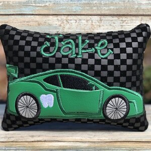 May include: A black and gray checkered pillow with the name "Jake" embroidered in green script. A green race car with a tooth graphic is also embroidered on the pillow. The pillow is rectangular.