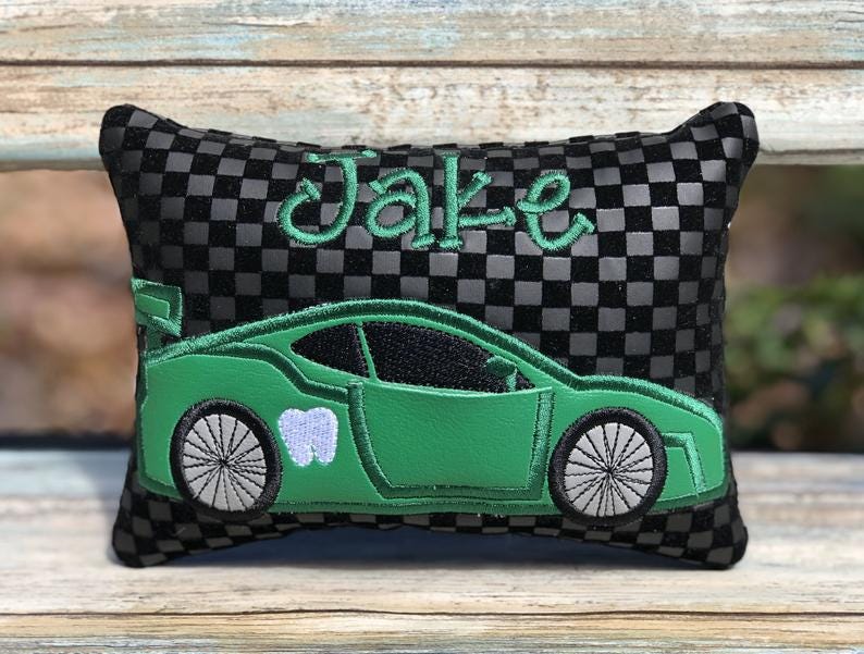 May include: A black and gray checkered pillow with the name "Jake" embroidered in green script. A green race car with a tooth graphic is also embroidered on the pillow. The pillow is rectangular.