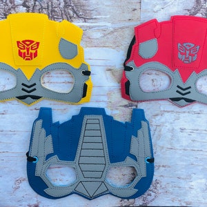 May include: Three felt masks of the Transformers characters, Optimus Prime, Bumblebee, and Megatron. The masks are yellow, blue, and red, respectively. Each mask has a gray face with eye holes and a black strap.