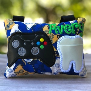 May include: A blue and yellow pillow with a black and green video game controller, a white tooth, and the name "Aver" embroidered on it.