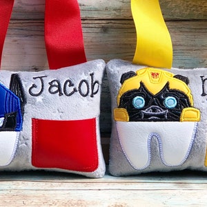 May include: Two gray tooth fairy pillows with red and yellow ribbon handles. One pillow features a blue and black robot design, the name "Jacob", and a red pocket. The other has a yellow and black robot design, the name "Mason", and a yellow pocket.