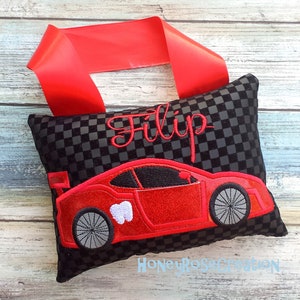 May include: A black and gray checkered tooth fairy pillow with a red race car design and the name "Filip" embroidered in red. A red ribbon is attached to the top of the pillow.