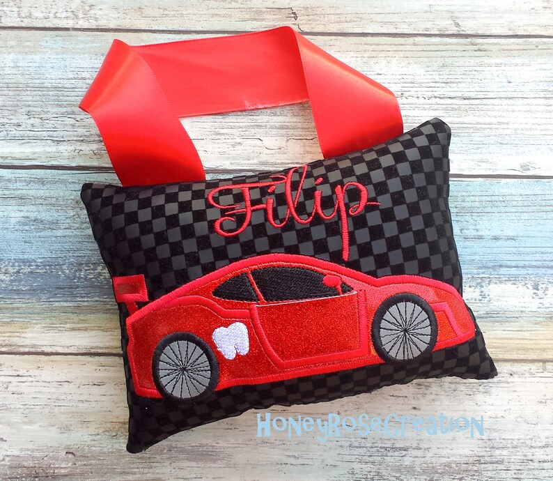 May include: A black and gray checkered tooth fairy pillow with a red race car design and the name "Filip" embroidered in red. A red ribbon is attached to the top of the pillow.