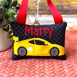 May include: Black and gray checkered tooth fairy pillow with a yellow race car design and the name "Matty" embroidered in red. Features a red ribbon for hanging.