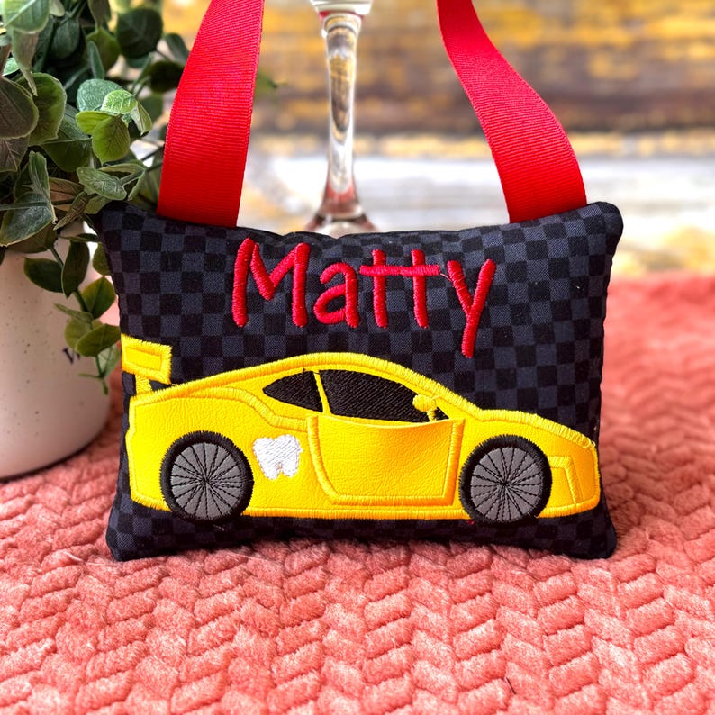 May include: Black and gray checkered tooth fairy pillow with a yellow race car design and the name "Matty" embroidered in red. Features a red ribbon for hanging.