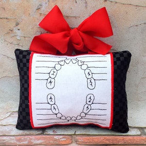 May include: A small, rectangular tooth fairy pillow with a black and white checkered pattern on the sides. The front features a white fabric with a red border and a diagram of teeth. A large red bow is attached at the top.