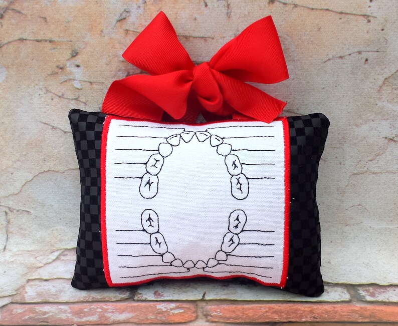 May include: A small, rectangular tooth fairy pillow with a black and white checkered pattern on the sides. The front features a white fabric with a red border and a diagram of teeth. A large red bow is attached at the top.