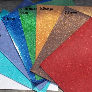 May include: A fan of glitter paper in various colors, including white, light blue, light purple, navy, Christmas green, orange, brown, and red. Each sheet is labeled with its color name.
