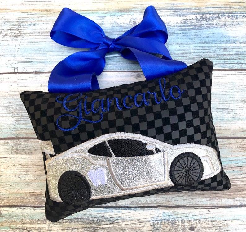 May include: A black and gray checkered pillow with a silver car design and the name "Giancarlo" embroidered in blue. A blue ribbon is tied at the top of the pillow.