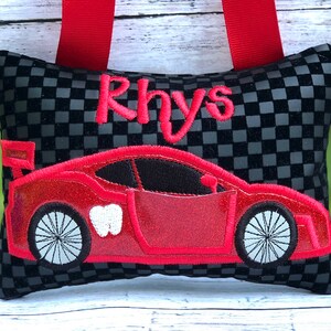 May include: A black and gray checkered tooth fairy pillow with a red race car design and the name "Rhys" embroidered in red. The pillow has red ribbon straps.