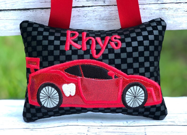 May include: A black and gray checkered tooth fairy pillow with a red race car design and the name "Rhys" embroidered in red. The pillow has red ribbon straps.