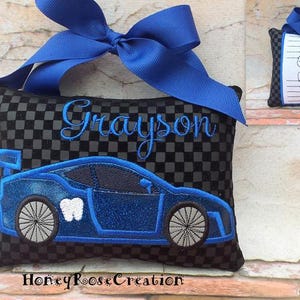 May include: A black and gray checkered tooth fairy pillow with a blue race car design and the name "Grayson" embroidered in blue. The pillow has a blue bow at the top. A smaller pillow with a tooth chart is also visible.