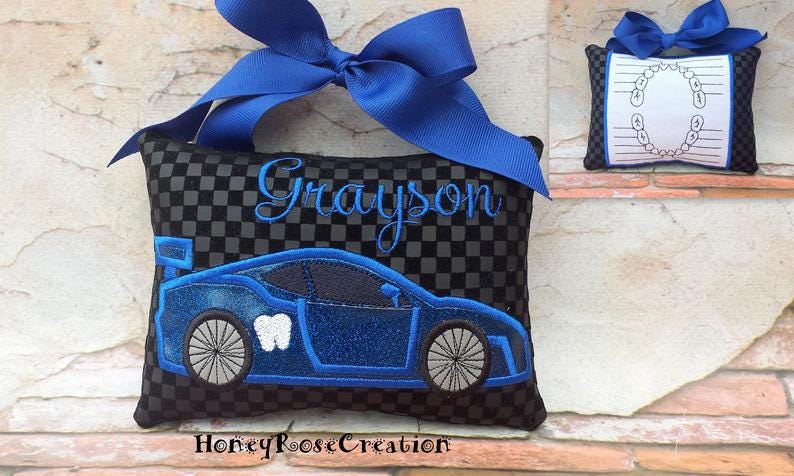 May include: A black and gray checkered tooth fairy pillow with a blue race car design and the name "Grayson" embroidered in blue. The pillow has a blue bow at the top. A smaller pillow with a tooth chart is also visible.