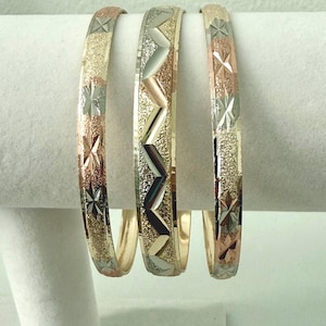 May include: Three metal bangles featuring a tri-color design of yellow, rose, and white gold tones. The bracelets display intricate textures, including diamond-cut star patterns and a central zigzag motif with alternating smooth and glittery finishes.