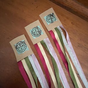 May include: Three handmade decorative streamers with a clock design. Each streamer features a cardboard base with a stamped clock image on a blue background. Colorful ribbons in shades of red, green, and purple cascade from the base, creating a whimsical effect.