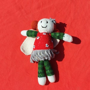 May include: A handmade knitted doll made with red, green and white yarn. The doll has a smiling face, a red dress with white flowers and white wings.