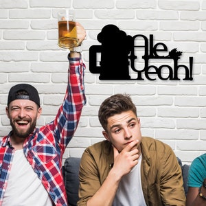 Może przedstawiać: Trzech przyjaciół siedzi na kanapie przed ceglaną ścianą. Jeden z nich trzyma kufel piwa i unosi go w górę. Na ścianie znajduje się czarny metalowy znak z napisem "Ale Yeah!" i sylwetką kufla piwa.