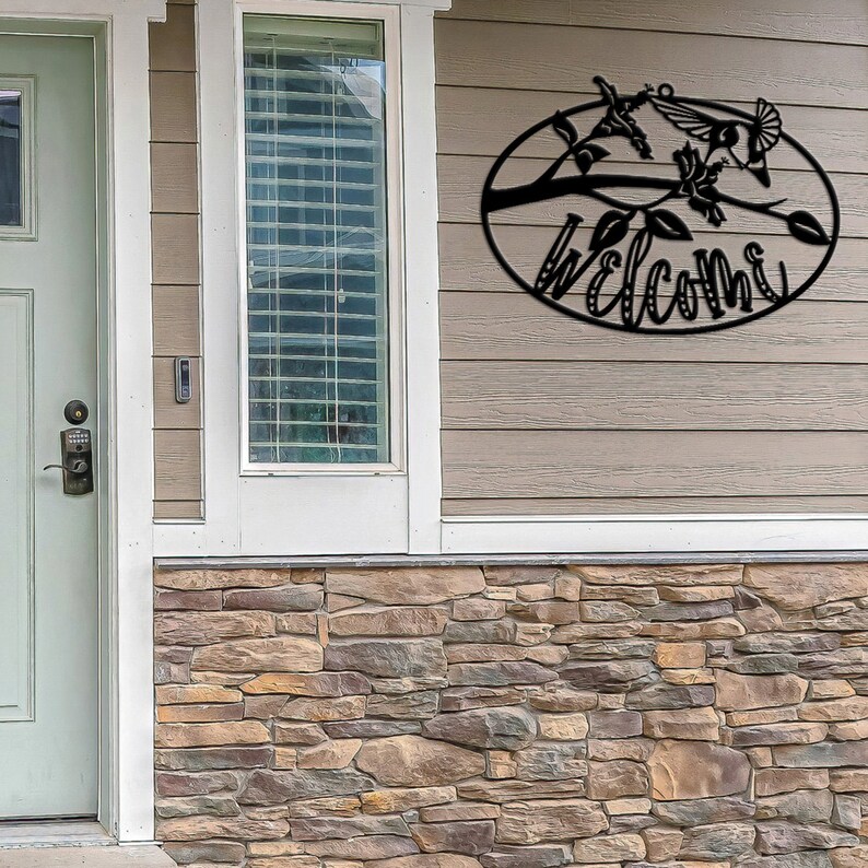 May include: Black metal welcome sign with a hummingbird and floral design. The sign is oval shaped and hangs on a light brown wooden wall.