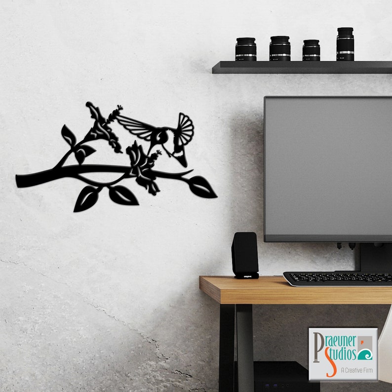 May include: Black metal wall art featuring a hummingbird in flight, perched on a branch with flowers. The art is mounted on a white wall, with a desk and computer monitor in the background.