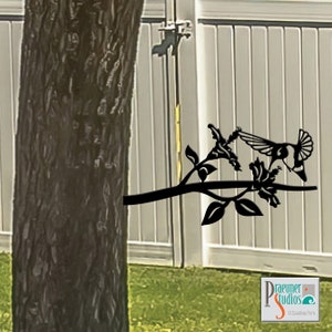 May include: Black metal hummingbird sculpture perched on a branch with flowers. The sculpture is attached to a tree trunk.