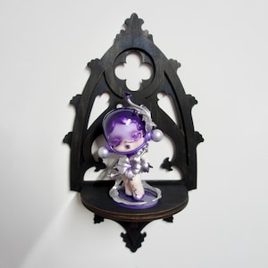 May include: A black gothic wooden shelf with a decorative cutout, displaying a purple figurine. The figurine features a helmet-shaped head and silver details. The shelf is mounted on a white wall, creating a contrast. The shelf's dimensions are approximately 20 cm.