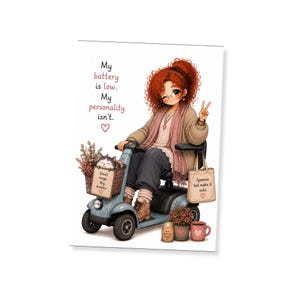 Op de afbeelding: Een wenskaartillustratie met een persoon met krullend rood haar op een lichtblauwe scootmobiel. De tekst luidt: My battery is low. My personality isn't. Een mandje bevat een kat en een bordje met de tekst Small range Big dreams.