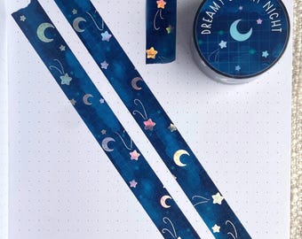 May include: Two rolls of blue washi tape with a moon and star pattern. The tape is on a white sheet of paper with a dotted pattern. The tape is labeled "Dreamy Starry Night".