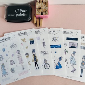 May include: Six white sticker sheets titled My Christian Girl featuring illustrations of people, bicycles, cassette tapes, and floral motifs in blue and yellow tones, accompanied by a black ink pad and a small box of wooden alphabet stamps.