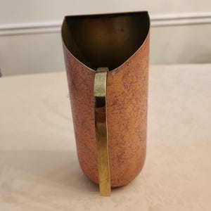 Puede incluir: Una jarra de metal color cobre con un asa de lat&oacute;n. La jarra tiene una superficie texturizada y una parte superior curvada. El asa es rectangular y est&aacute; unida al lateral de la jarra.