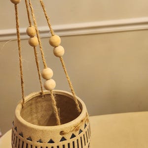 May include: A beige ceramic hanging planter with a geometric design of vertical lines and diamond shapes. The planter is suspended by jute rope with wooden beads. The interior is a darker beige, and the bottom has a light blue hue.