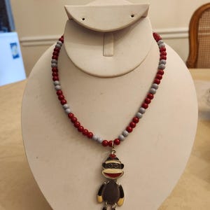 May include: A beaded necklace with a sock monkey pendant. The necklace features alternating red and gray beads. The pendant is a sock monkey with a red hat, black and yellow feet, and a smiling face. The necklace is displayed on a white bust.