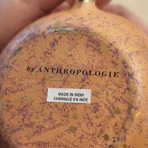 Puede incluir: Primer plano de un objeto decorativo naranja y dorado con una superficie texturizada. El texto "by ANTHROPOLOGIE" es visible, junto con una etiqueta que dice "MADE IN INDIA FABRIQU&Eacute; EN INDE".
