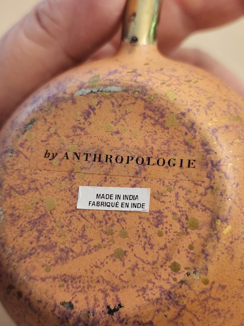 Puede incluir: Primer plano de un objeto decorativo naranja y dorado con una superficie texturizada. El texto "by ANTHROPOLOGIE" es visible, junto con una etiqueta que dice "MADE IN INDIA FABRIQU&Eacute; EN INDE".