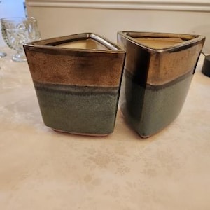 May include: Two ceramic planters with a triangular shape. The top section is brown, the middle is a muted green, and the interior is beige. The planters are on a light-colored surface.