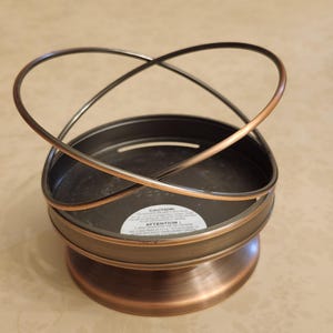 May include: A round metal candle holder with a brushed bronze finish. The base is circular with a flared bottom, and the top features two intersecting curved metal bands that form an open, orbital design. A white caution label is visible inside.