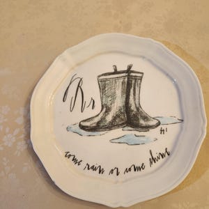 Pu&ograve; includere: Un piatto in ceramica bianca con bordo smerlato presenta un'illustrazione disegnata a mano di stivali da pioggia in bianco e nero. Il piatto include il testo "Rr" e "come rain or come shine" in scrittura nera.