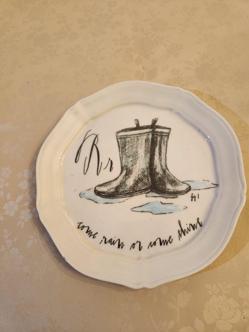 Pu&ograve; includere: Un piatto in ceramica bianca con bordo smerlato presenta un'illustrazione disegnata a mano di stivali da pioggia in bianco e nero. Il piatto include il testo "Rr" e "come rain or come shine" in scrittura nera.