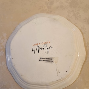 Pu&ograve; includere: Piatto in ceramica bianca con bordo smerlato. Il piatto presenta la scritta "LINEA CARTA by Dra Pyari" e un adesivo con la scritta "Dishwasher & Microwave Safe Made in China/Fabriqu&eacute; en Chine."