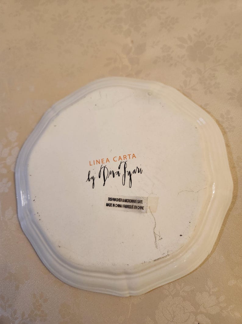 Pu&ograve; includere: Piatto in ceramica bianca con bordo smerlato. Il piatto presenta la scritta "LINEA CARTA by Dra Pyari" e un adesivo con la scritta "Dishwasher & Microwave Safe Made in China/Fabriqu&eacute; en Chine."