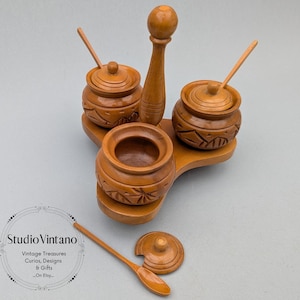 May include: A wooden condiment set with three small pots, lids, and spoons. The pots are carved with a floral design and sit on a three-leaf-shaped base. The set includes a tall, central piece. The color is a warm, light brown.