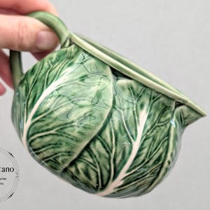May include: A ceramic pitcher shaped like a green cabbage head with a glossy finish and visible crackle glaze texture. The pitcher features a curved handle and a spout, with embossed leaf veins in shades of dark and light green throughout.
