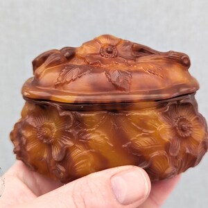 May include: A small, oval-shaped, brown and amber glass trinket box with a floral design. The box has a hinged lid and is decorated with raised flowers and leaves.