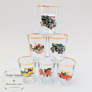 May include: Six clear shot glasses with gold rims, each decorated with a different vintage car design in various colours. The glasses are stacked in a pyramid shape against a white background.
