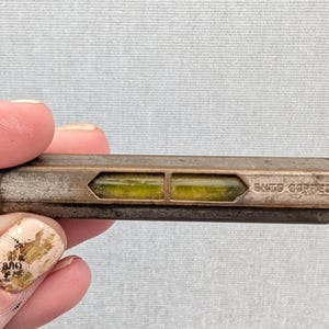 Puede incluir: Una antigua herramienta de nivel con forma rectangular y dos viales llenos de líquido verde. La carcasa metálica tiene un aspecto deslustrado, con el texto "GNTS CORRECT" visible en el lateral. La herramienta está sujeta por una persona.